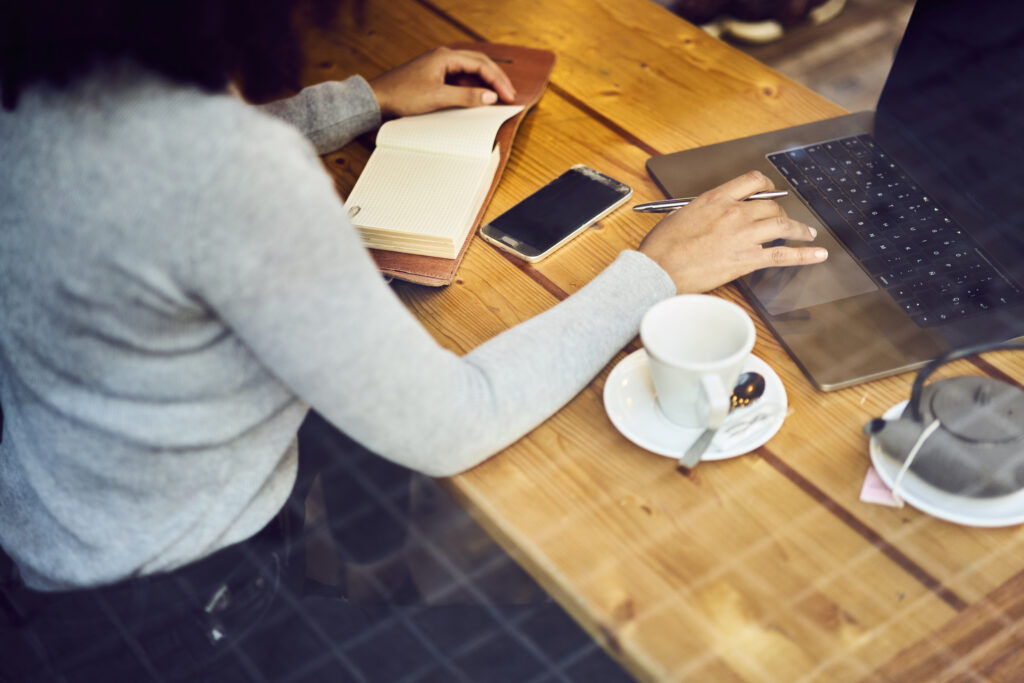 Person writing notes at a wooden desk with a laptop, phone and coffee
