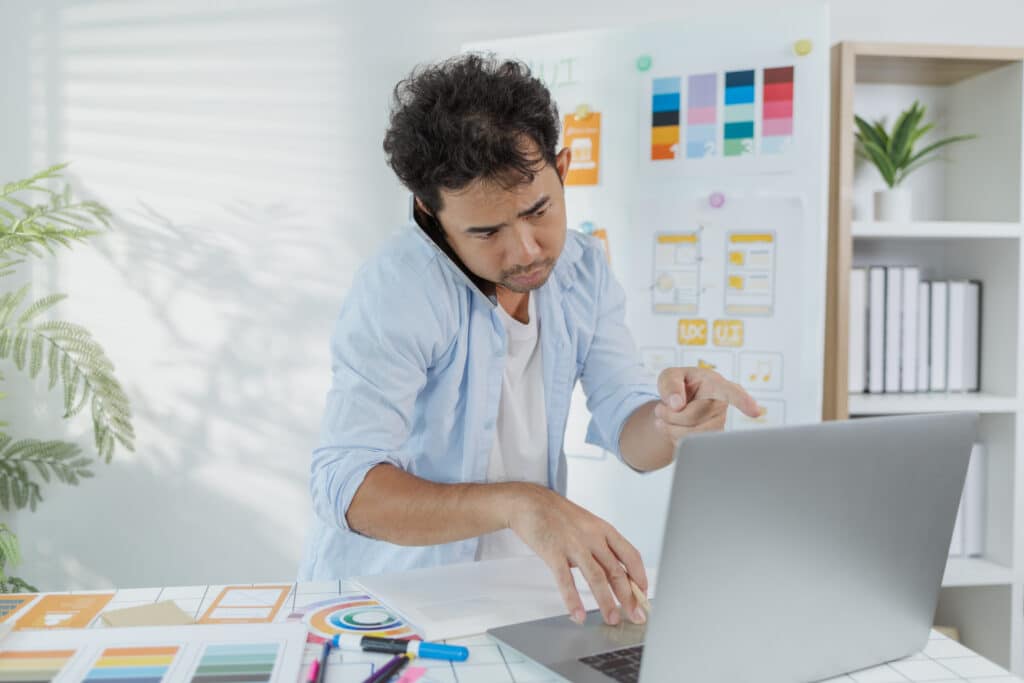 Designer on a phone call at a desk with a laptop and colour swatches on the wall